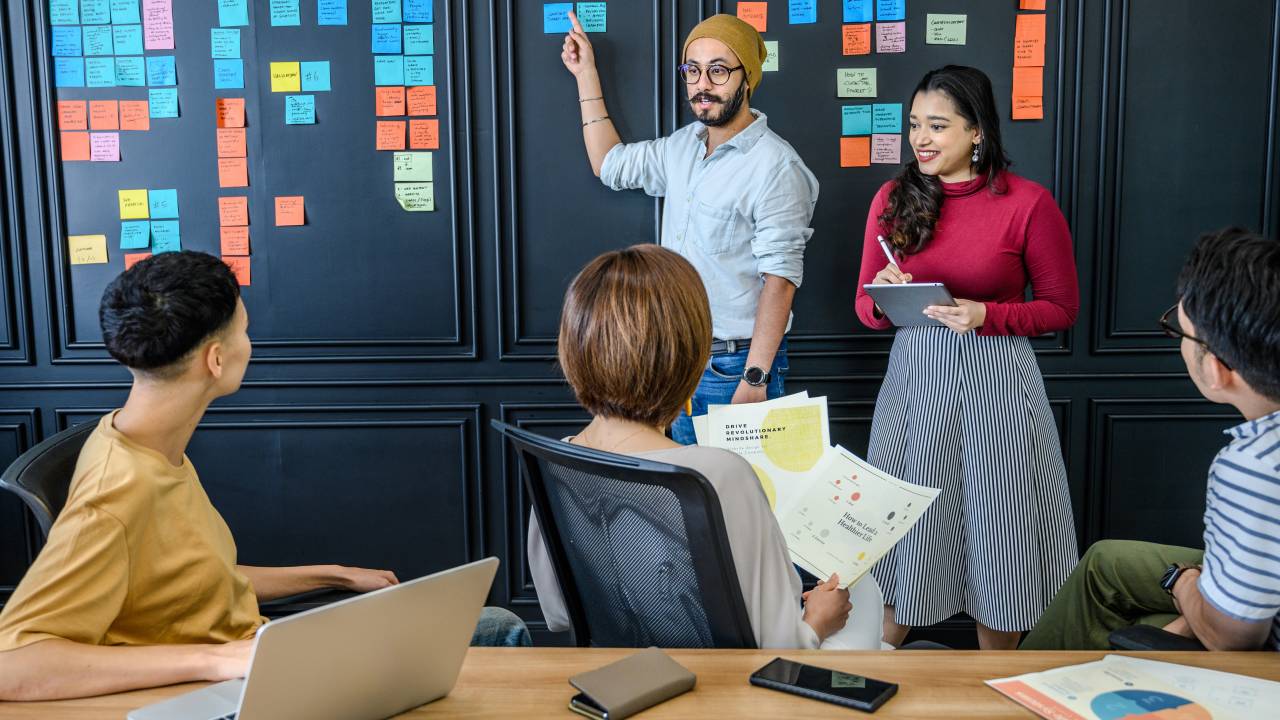 Homem aponta uma parede com papeis coloridos colados, em uma reunião de trabalho, enquanto os demais prestam atenção