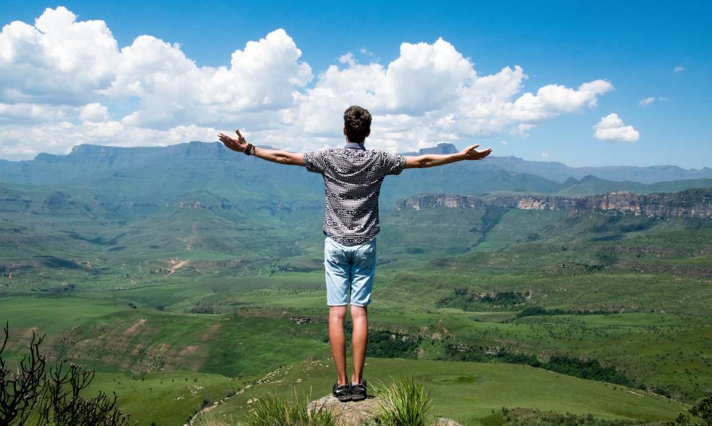 Um homem está no pico de uma montanha, de braços abertos, admirando a vista.