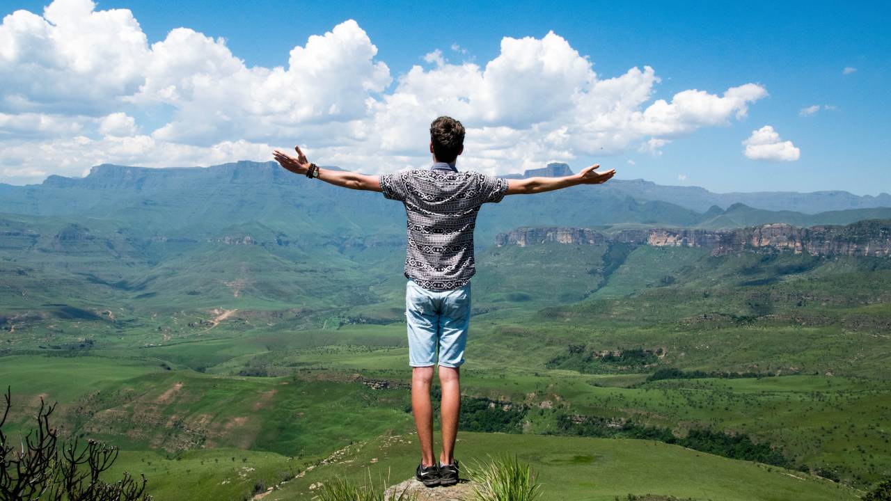 Um homem está no pico de uma montanha, de braços abertos, admirando a vista.
