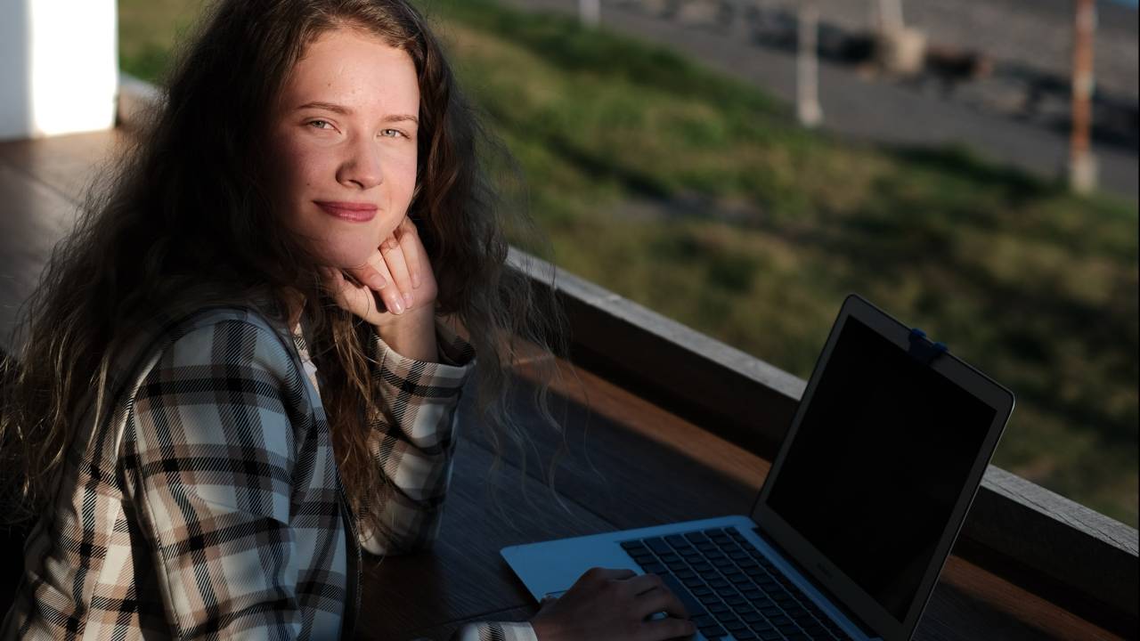 Uma mulher está vestida com uma camisa xadrez enquanto mexe em um notebook. Ela está de frente para uma janela grande cuja vista é uma praia