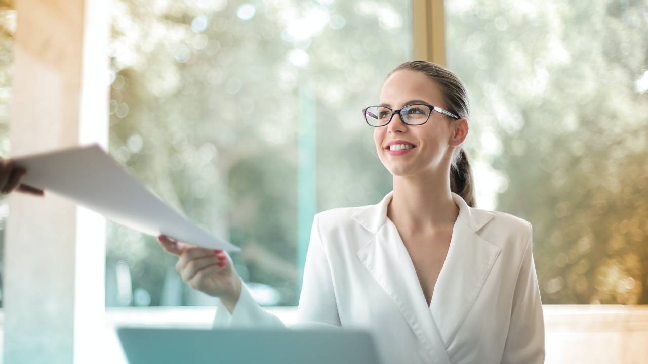 Uma mulher está sentada em frente a um computador. Ela veste um blazer branco e sorri enquanto parece entregar uma folha de papel a alguém