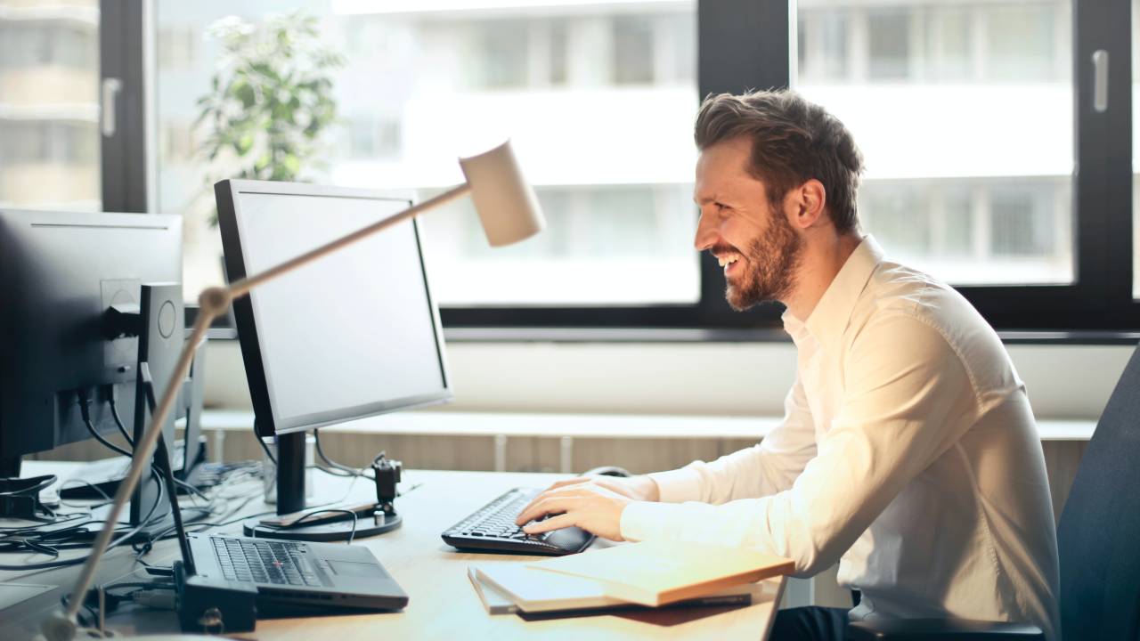 Um homem vestido socialmente está sentado em uma mesa enquanto olha para a tela do computador e sorri.