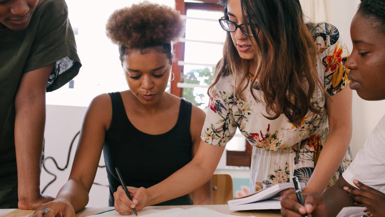 Quatro mulheres estão em volta de uma mesa. Duas delas estão sentadas, as outras duas debruçadas. Todas parecem analisar papéis.