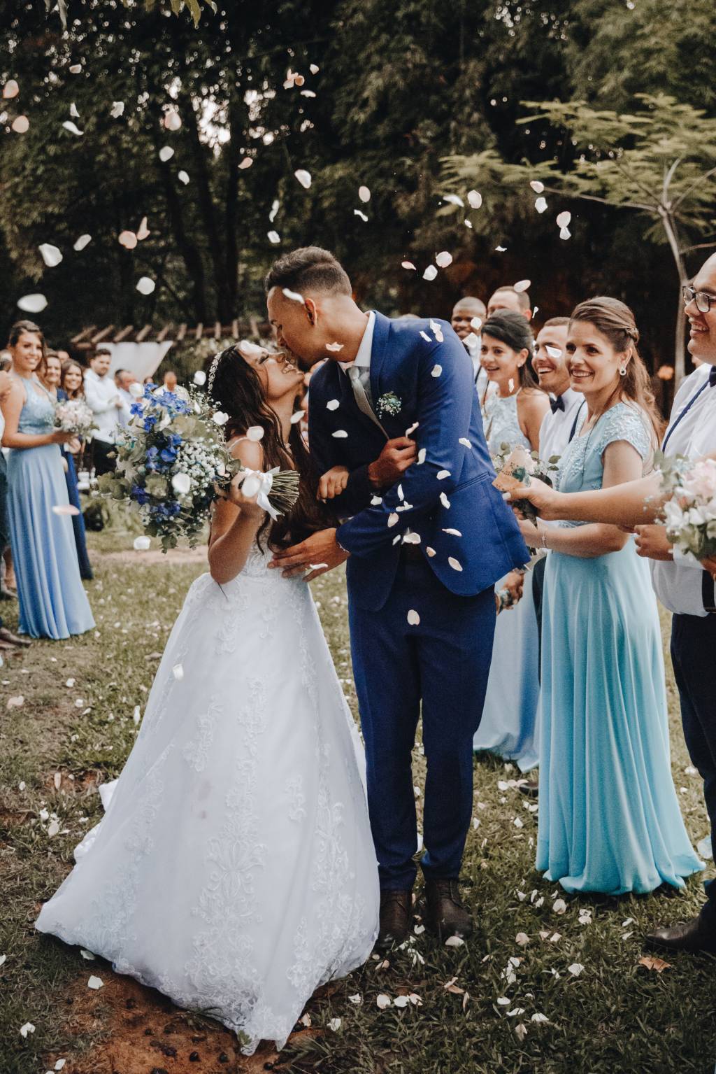 Noivos se beijam em casamento no jardim, com chuva de papel picado e convidados ao redor. madrinhas vestem tons de azul claro