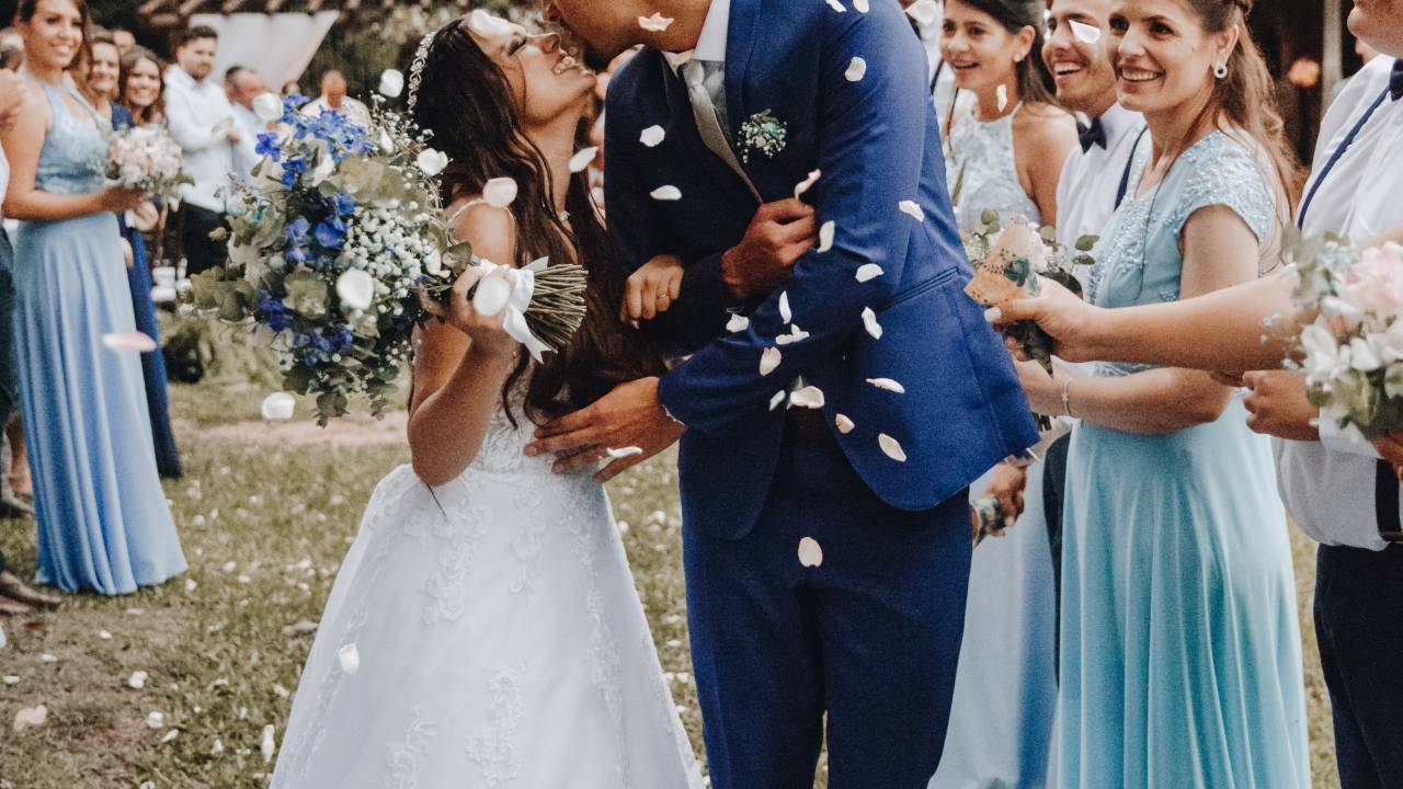 Noivos se beijam em casamento no jardim, com chuva de papel picado e convidados ao redor. madrinhas vestem tons de azul claro