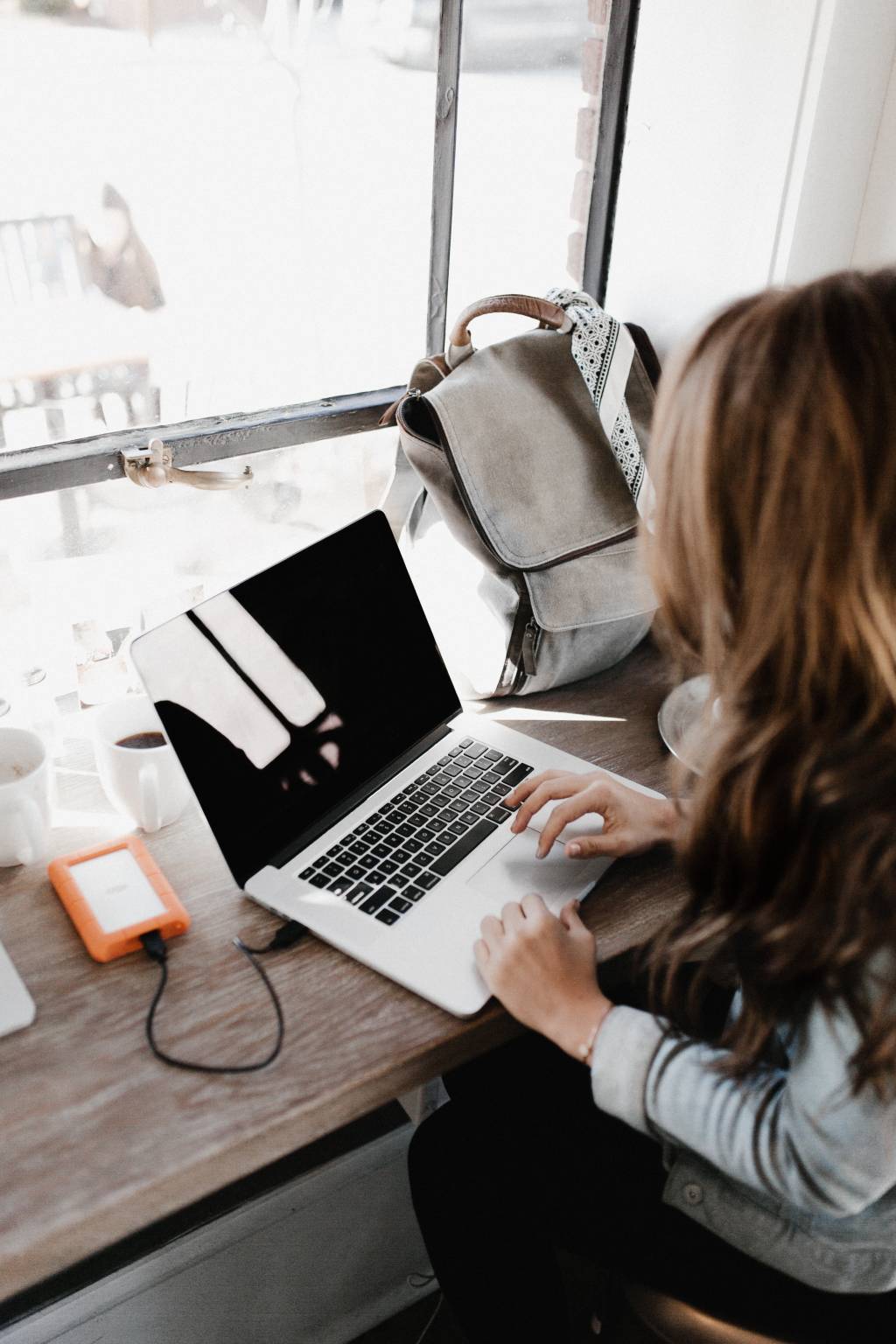 Uma mulher está trabalhando em um notebook. Sua mochila está na mesa, ao lado do computador.