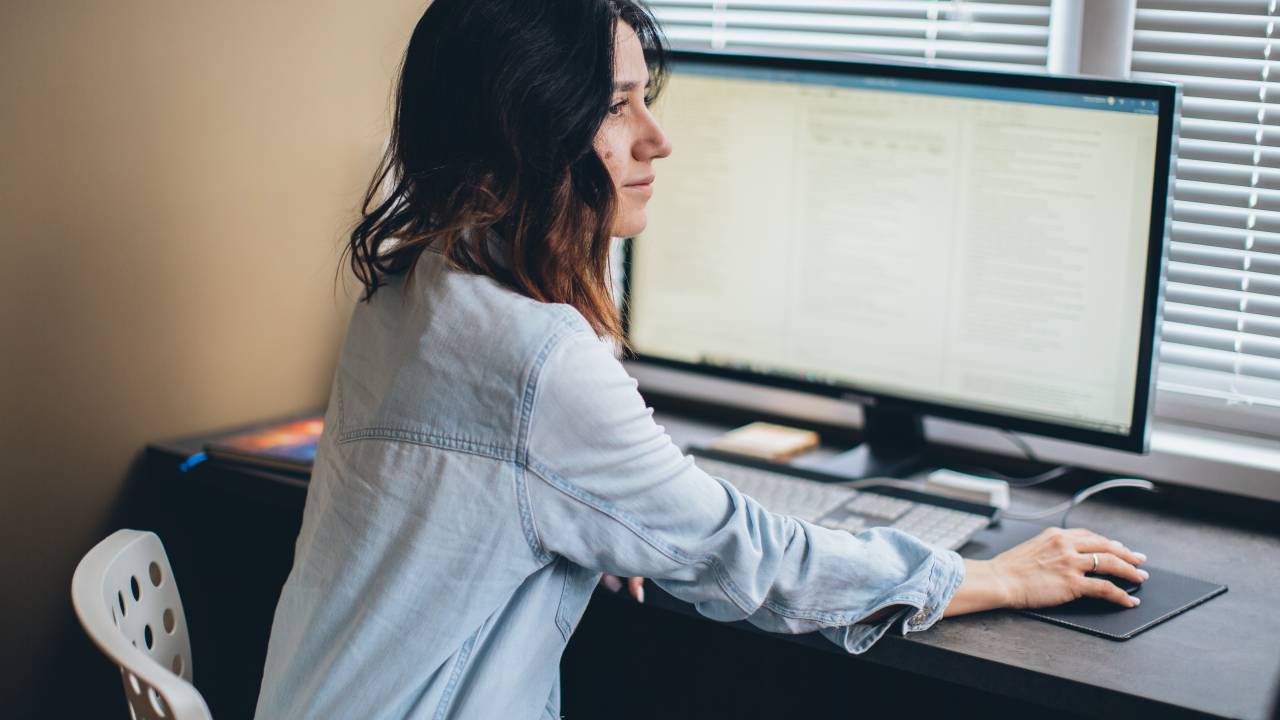Uma mulher veste uma camisa jeans, está sentada em frente a um computador e olha para o lado