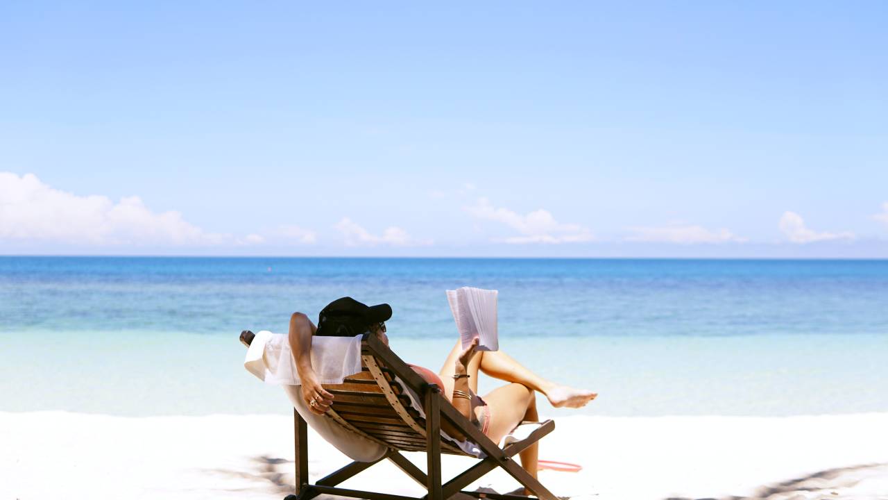 Mulher lê livro na praia sentada em uma espreguiçadeira de madeira diante de um mar turquesa