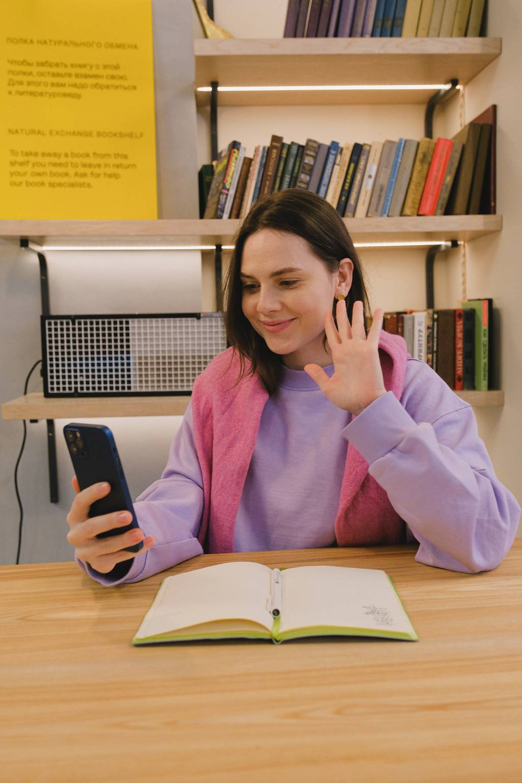 Uma jovem está sentada em frente a uma mesa com um caderno aberto. Ela interage por vídeo com alguém por meio de um smartphone