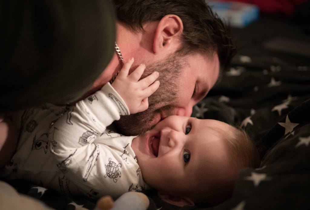 Homem com barba por fazer e corrente no pescoço beija bebê sorridente