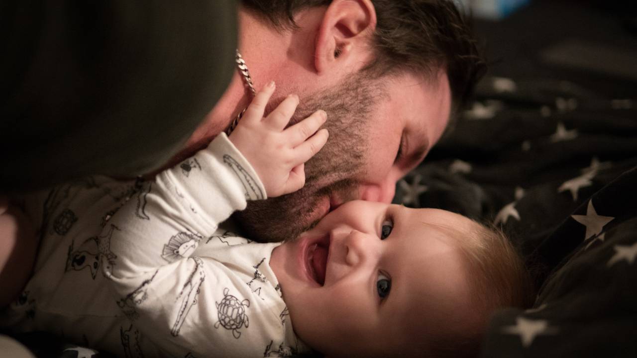 Homem com barba por fazer e corrente no pescoço beija bebê sorridente