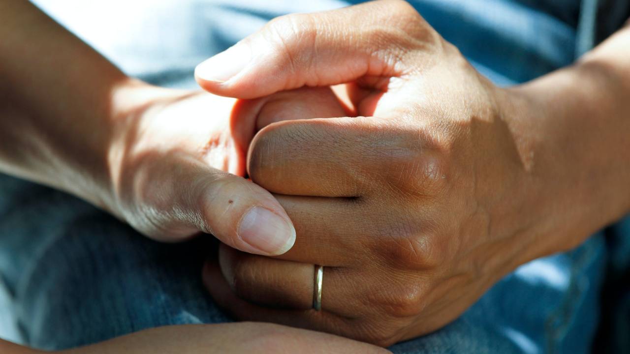A imagem mostra as mãos de um casal entrelaçadas em um ambiente que parece ser um hospital