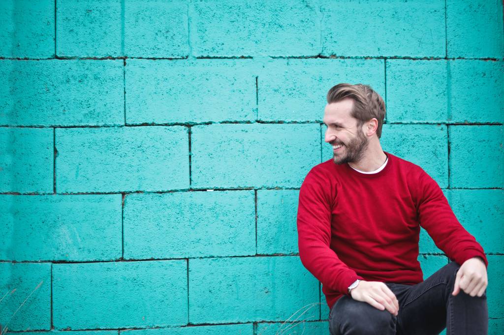 Homem com barba veste suéter vermelho e calça grafite e está encostado em uma parede azul turquesa