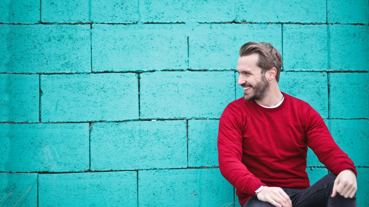Homem com barba veste suéter vermelho e calça grafite e está encostado em uma parede azul turquesa