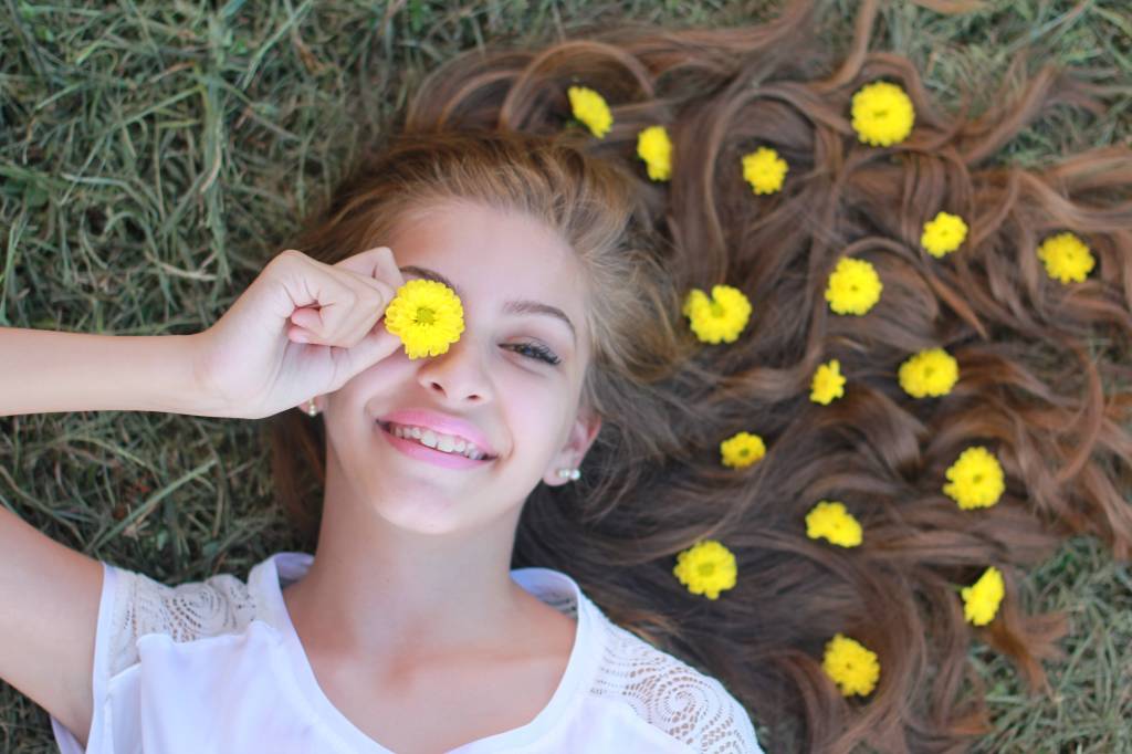 Mulher de cabelo ondulado loiro com nuances avermelhadas está deitada na grama, com flores amarelas nos fios e sorrindo para a câmera