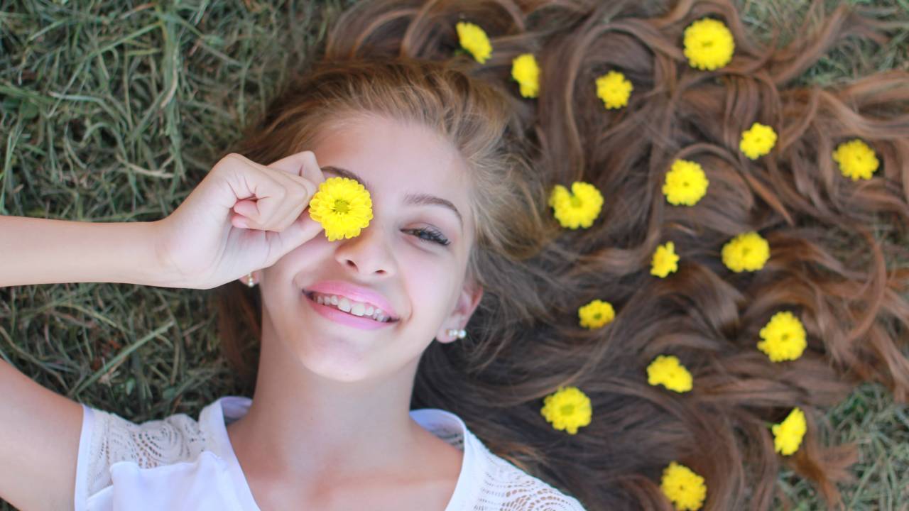 Mulher de cabelo ondulado loiro com nuances avermelhadas está deitada na grama, com flores amarelas nos fios e sorrindo para a câmera