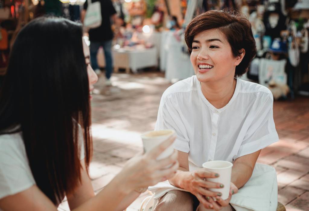 Mulheres tomam café ao ar livre e sorriem uma para a outra