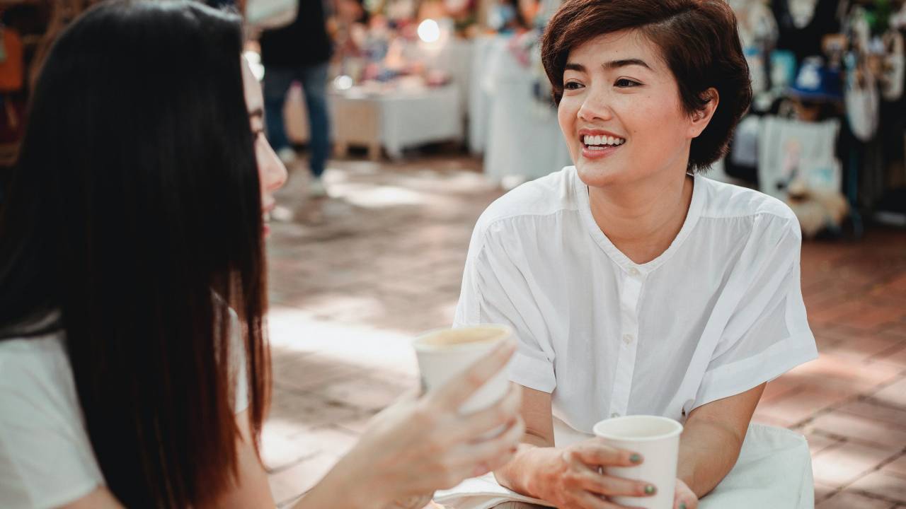 Mulheres tomam café ao ar livre e sorriem uma para a outra