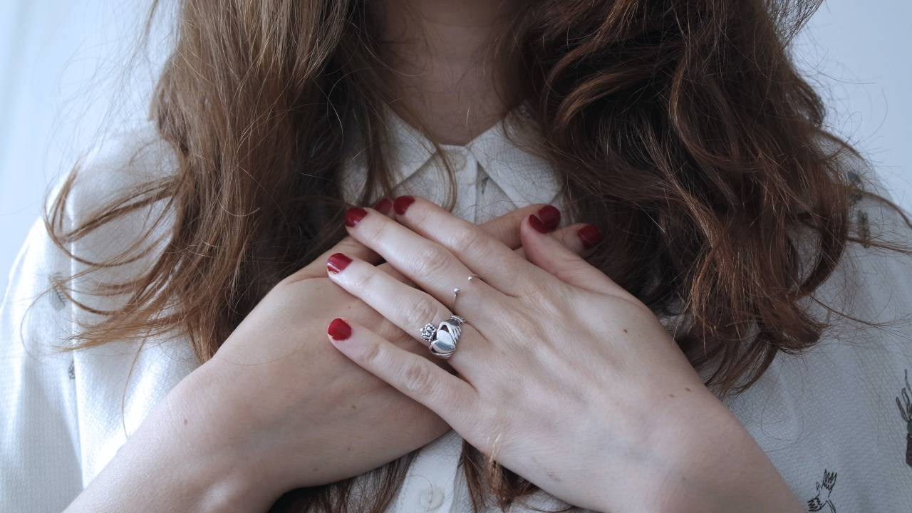Imagem mostra mãos de mulher sobre a região do peito formando um coração. Ela veste camisa branca, está com aneis e tem as unhas pintadas de vermelho