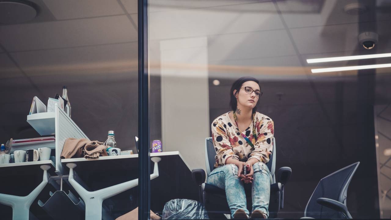 Mulher com calça jeans e blusa estampada está na cadeira do escritório olhando de forma contemplativa pela janela envidraçada do prédio, aparentando estar descontente com o trabalho