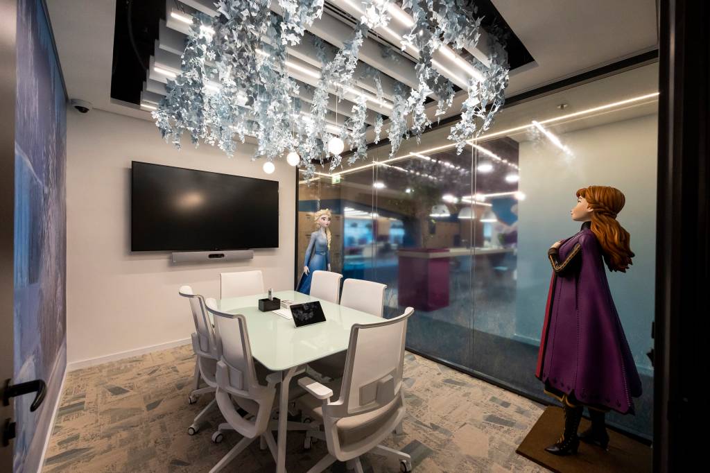 Disney frozen A sala de reunião tem as personagens Anna e Elza em tamanho rea, uma mesa com cadeiras e, no teto, flocos de neve
