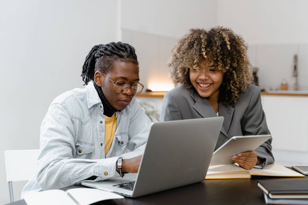 Dois profissionais negros estão sorrindo enquanto olham para a tela de um computador. Os dois estão sentados em uma mesa de escritório