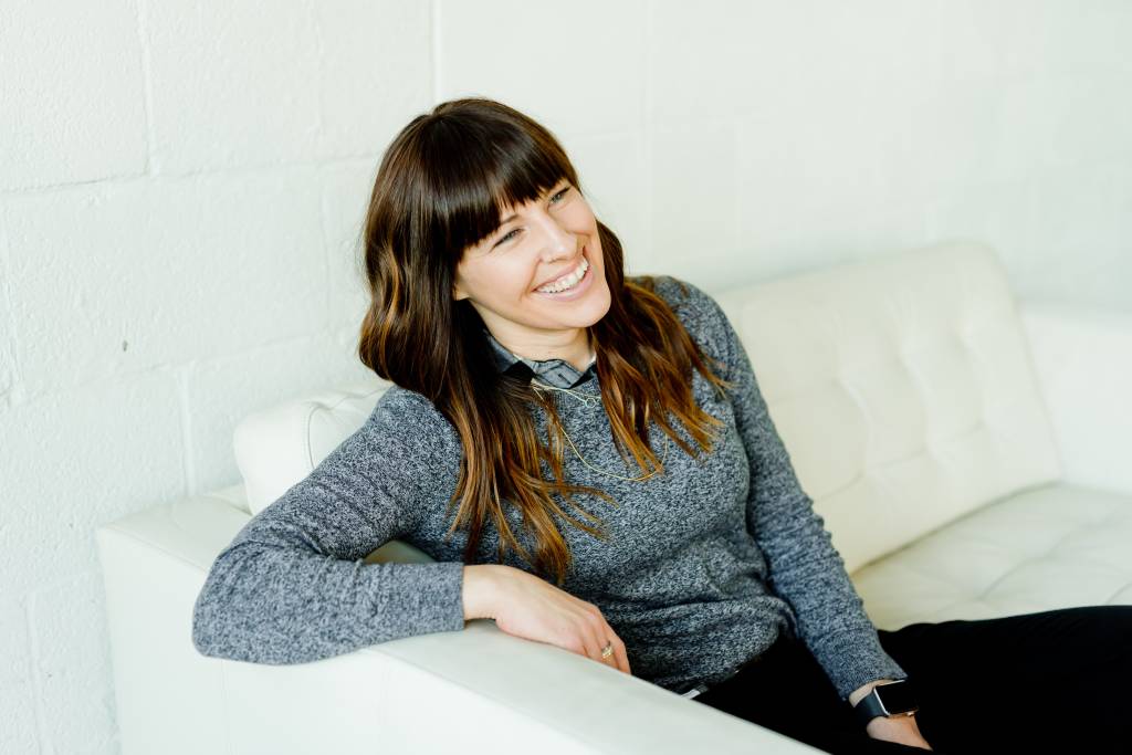 Mulher de cabelo comprido e franja vestindo suéter cinza está sentada ao sofá, sorrindo