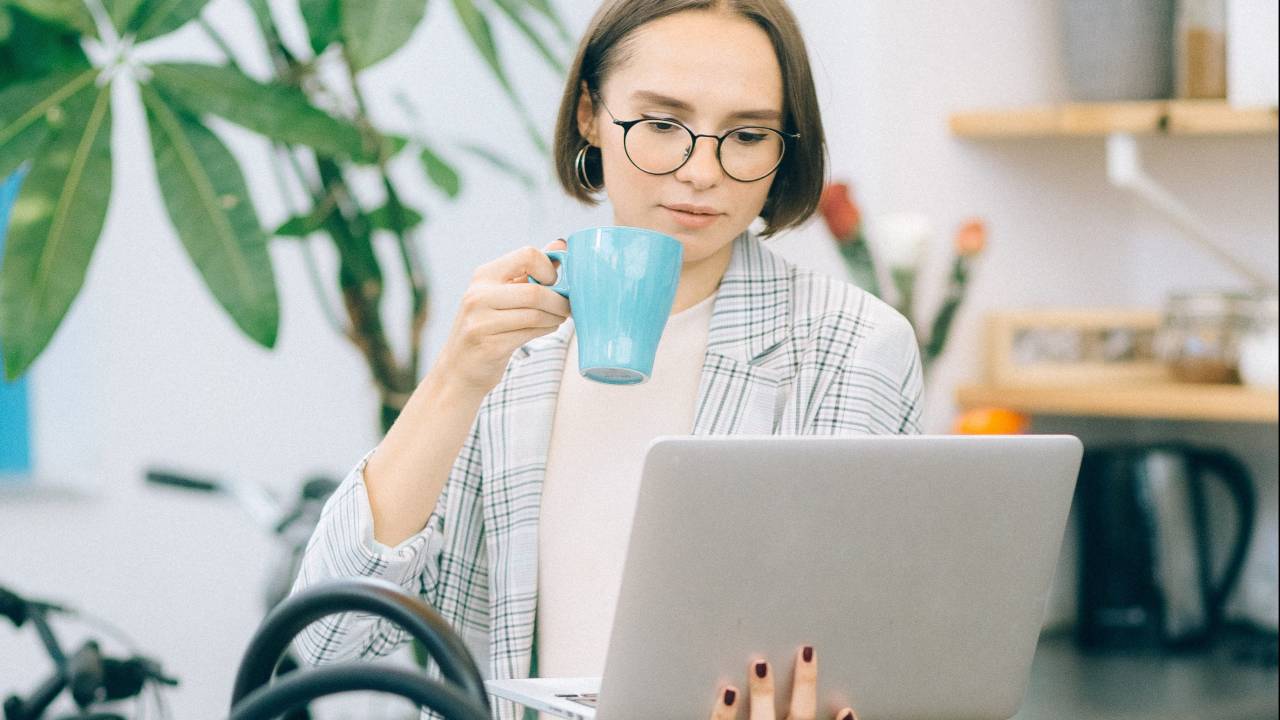 Uma mulher está na cozinha, bebendo algo em uma xícara azul enquanto segura o computador