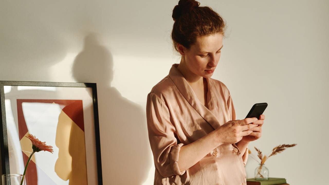 Uma mulher está apoiada a um móvel de sua casa, vestida com um pijama nude de manga comprida, enquanto vê o celular