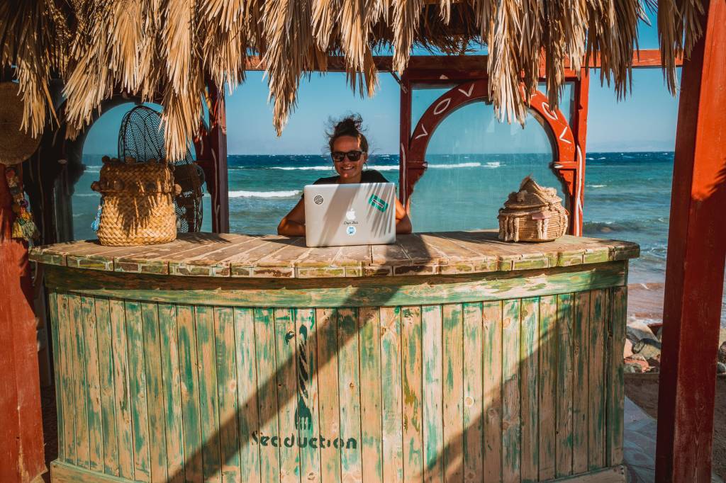 Mulher nômade digital trabalha com um notebook em uma praia. Ela está em um quiosque