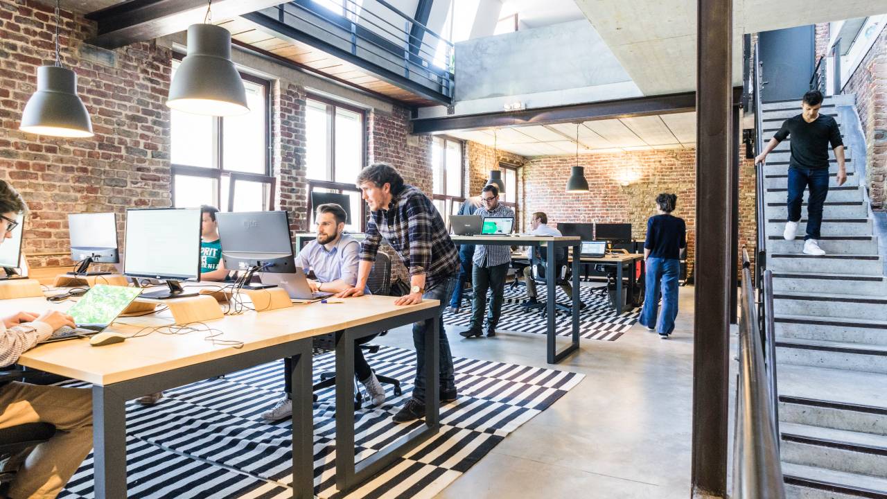 Imagem mostra funcionários de uma empresa trabalhando em um escritório com paredes de tijolinhos e muita luz natural