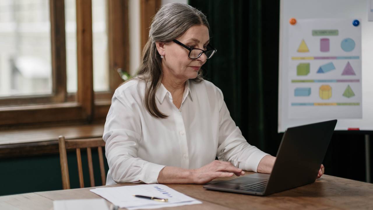Uma mulher de cabelos grisalhos e óculos está concentrada olhando para a tela de um computador