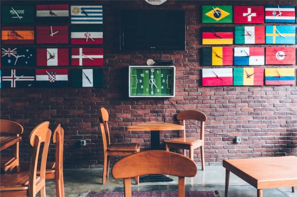 Um bar tem bandeiras de vários países fixadas em uma parede de tijolos. As mesas são de madeira e há um relógio em formato de campo de futebol na parede.