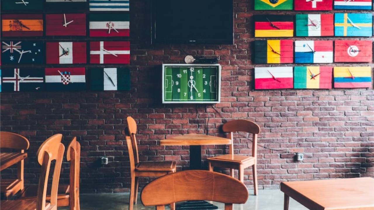 Um bar tem bandeiras de vários países fixadas em uma parede de tijolos. As mesas são de madeira e há um relógio em formato de campo de futebol na parede.