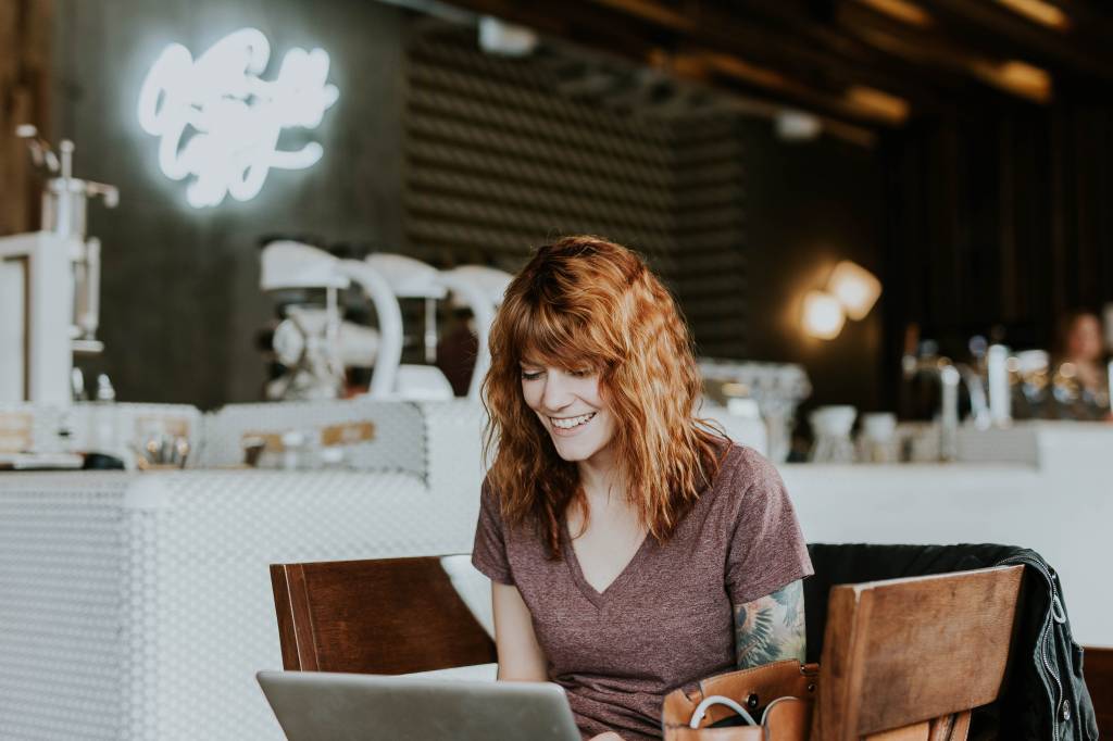 Uma mulher ruiva está sentada sorrindo em frente a um computador. Ela parece estar em uma cafeteria