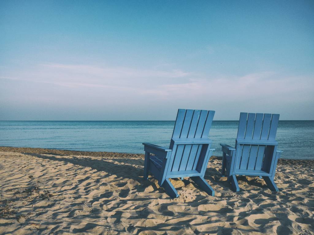 Duas cadeiras de madeira estão uma ao lado da outra sobre a areia de uma praia ensolarada e deserta