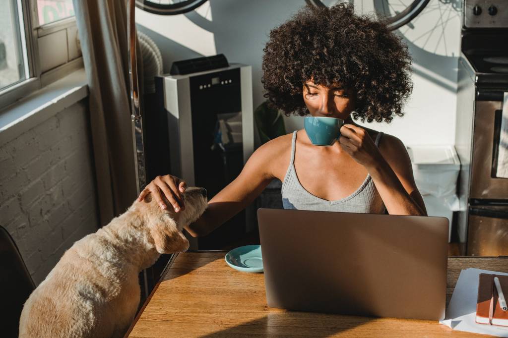Uma mulher está tomando uma bebida em uma xícara enquanto acaricia seu cachorro e trabalha no computador