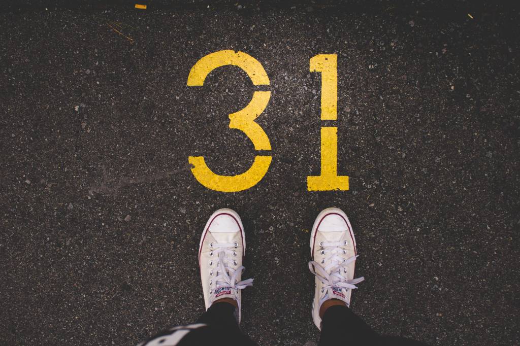 Imagem mostra uma pessoa em pé sobre o asfalto, com all star branco. No asfalto está pintado o número 31