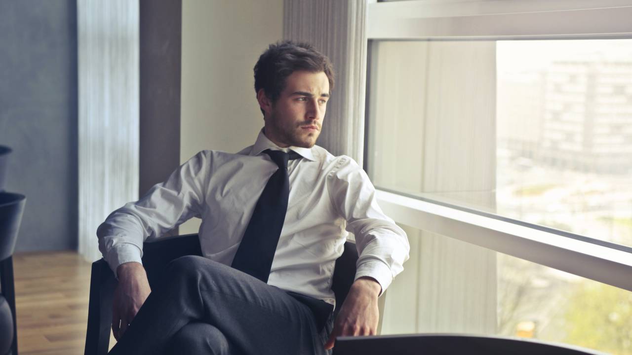 Homem executivo de camisa branca e gravata está sentado na cadeira do escritório olhando pela janela