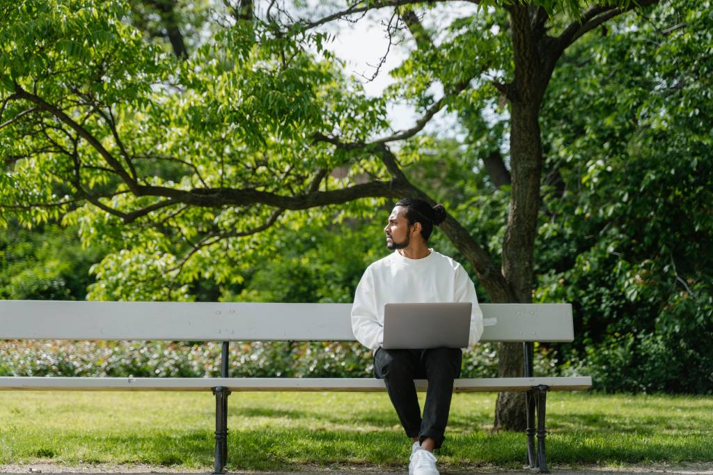 Homem com notebook sentado no banco de uma praça olha para o lado