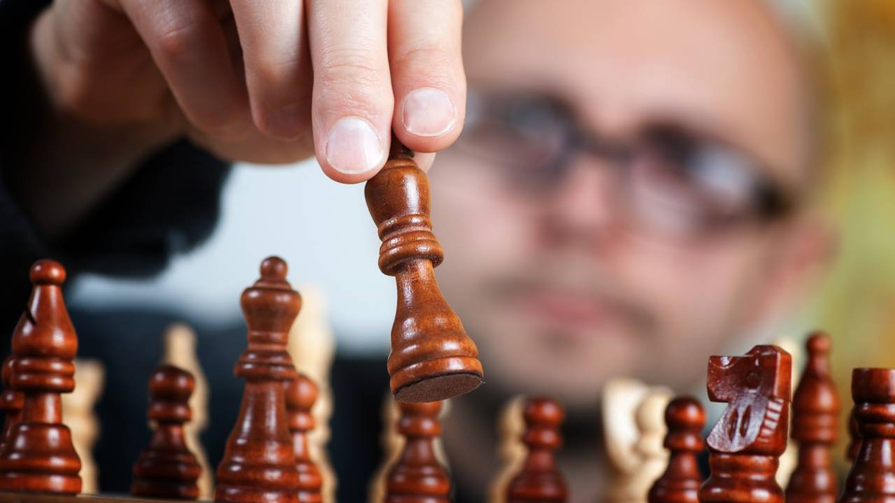 Um homem está movimentando peças de um xadrez. O primeiro plano mostra as peças sobre o tabuleiro e a mão do homem, que tem seu rosto em segundo plano desfocado