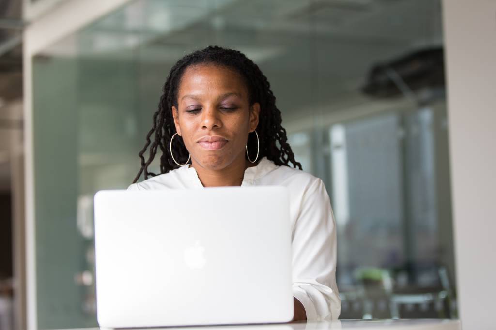 Uma mulher negra está atrás de um computador. Ela está com os lábios cerrados, vestida de branco, prestando atenção na tela