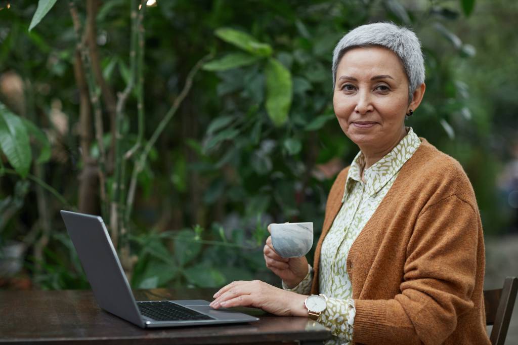Mulher de cabelo curto branco toma uma xícara de chá e trabalha em um jardim com um notebook