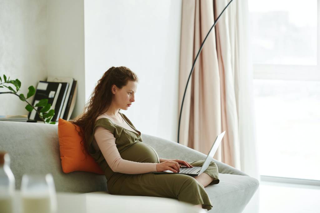 Uma mulher grávida esta sentada confortavelmente em um sofá de uma sala de estar enquanto trabalha com um computador no colo