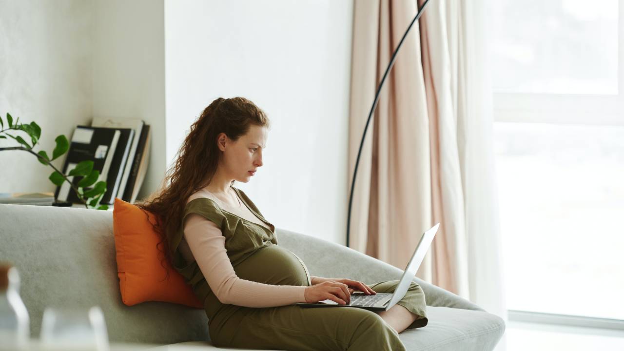 Uma mulher grávida esta sentada confortavelmente em um sofá de uma sala de estar enquanto trabalha com um computador no colo