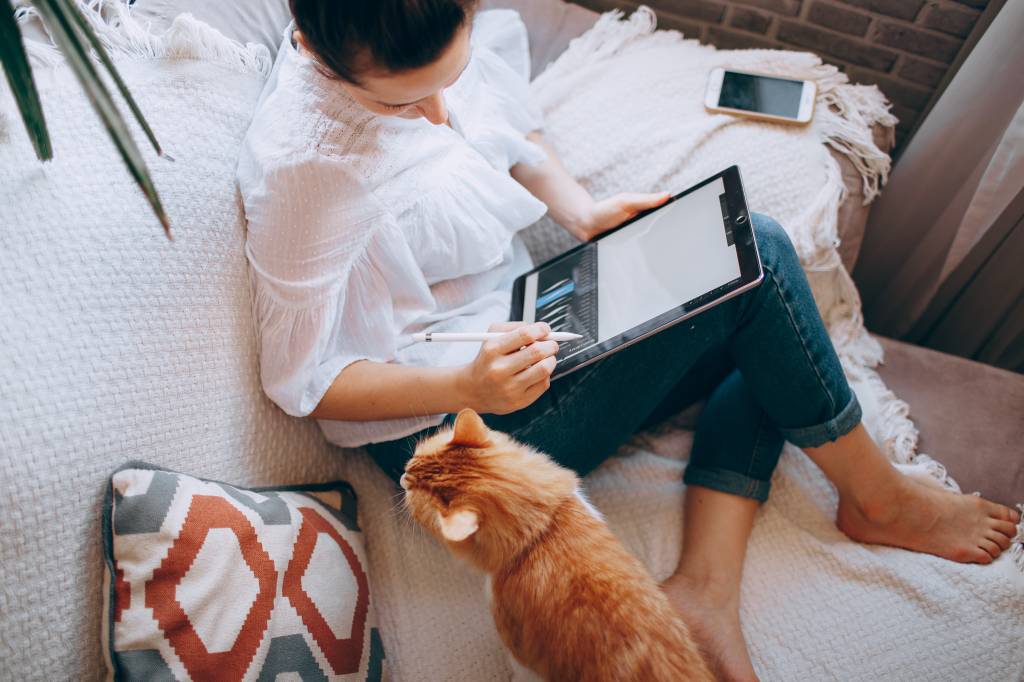 Mulher trabalha no sofá de casa com um tablet e um gato laranja ao lado