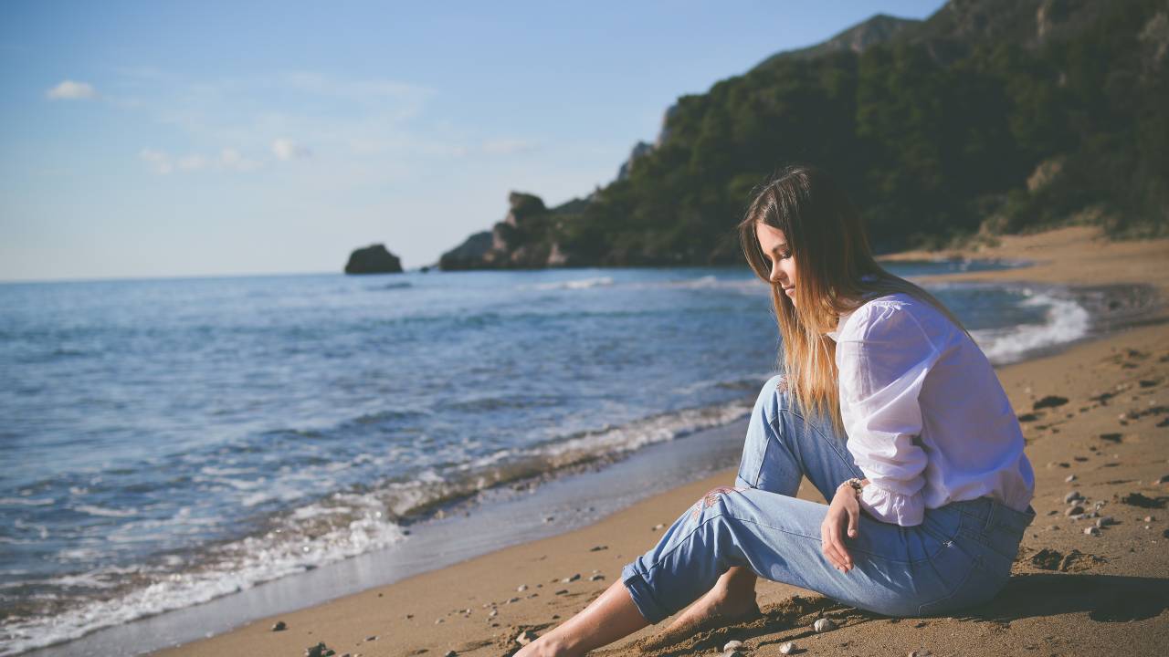 Mulher com calça azul claro e blusa branca está sentada na areia com os pés descalços