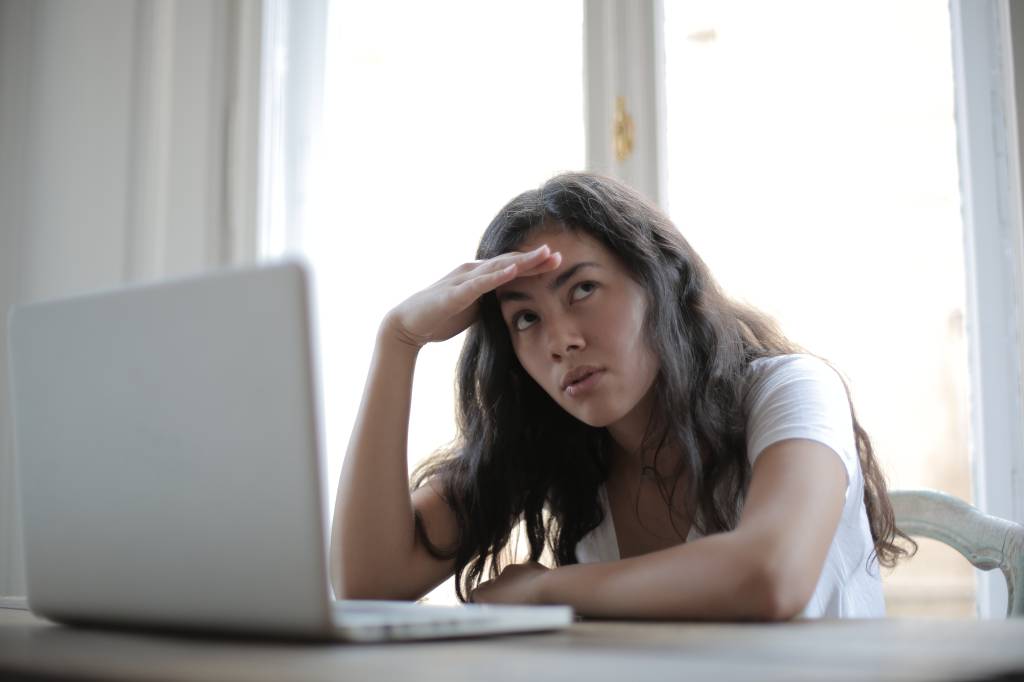 Uma mulher está sentada sob uma mesa em frente a um computador. Ela coloca uma das mãos na testa e parece preocupada