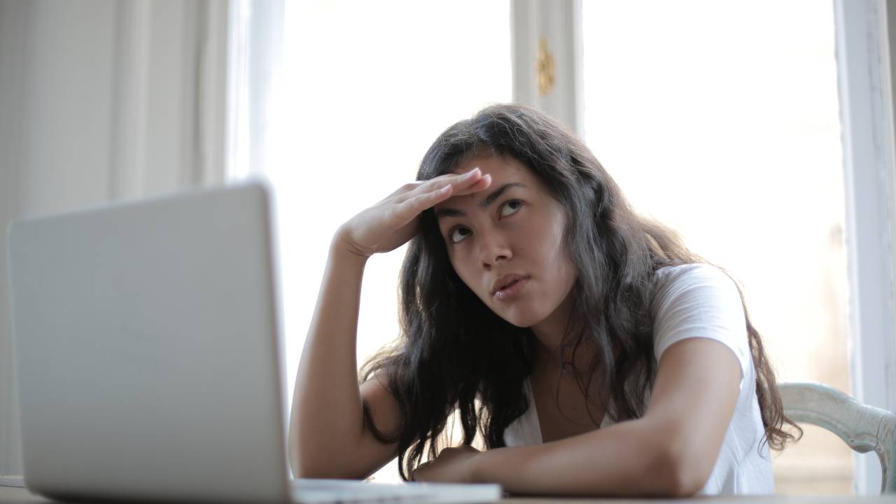 Uma mulher está sentada sob uma mesa em frente a um computador. Ela coloca uma das mãos na testa e parece preocupada