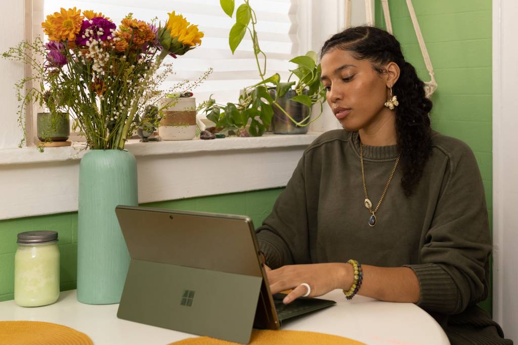 Uma mulher está sentada em frente a uma mesa aparentemente de uma cozinha, mexendo em seu tablet. Ela está perto de uma janela que tem plantas e flores
